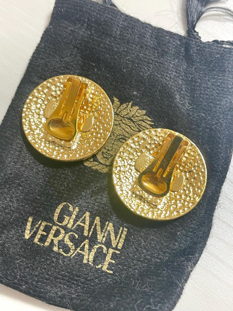 May include: A pair of gold-tone clip-on earrings with a textured circular design. The earrings are in a black velvet pouch with the text "GIANNI VERSACE" printed on it.
