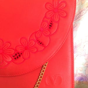 May include: A bright red leather clutch with a gold chain strap and a floral design. The clutch has a flap closure and the initials "VD" embossed on the front.
