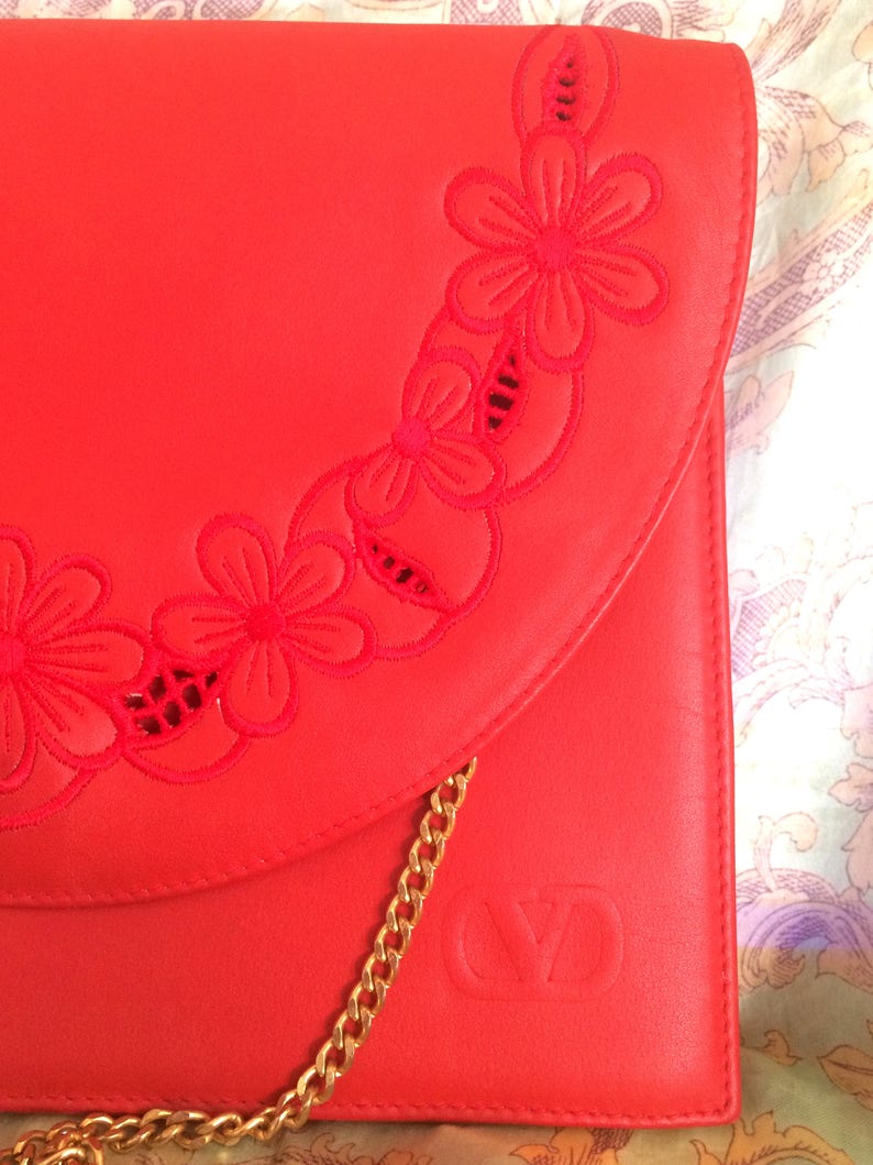 May include: A bright red leather clutch with a gold chain strap and a floral design. The clutch has a flap closure and the initials "VD" embossed on the front.