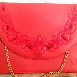 May include: A red leather clutch with a gold chain strap and a floral design. The floral design is cut out of the leather and is in a crescent shape.