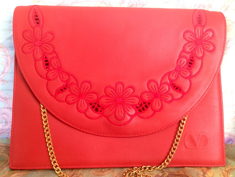 May include: A red leather clutch with a gold chain strap and a floral design. The floral design is cut out of the leather and is in a crescent shape.