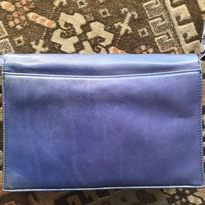May include: A rectangular, periwinkle leather handbag with a shoulder strap. The bag features a flap closure and a subtle texture. The strap is attached with silver-toned hardware. The bag is set against a patterned, neutral-toned background.