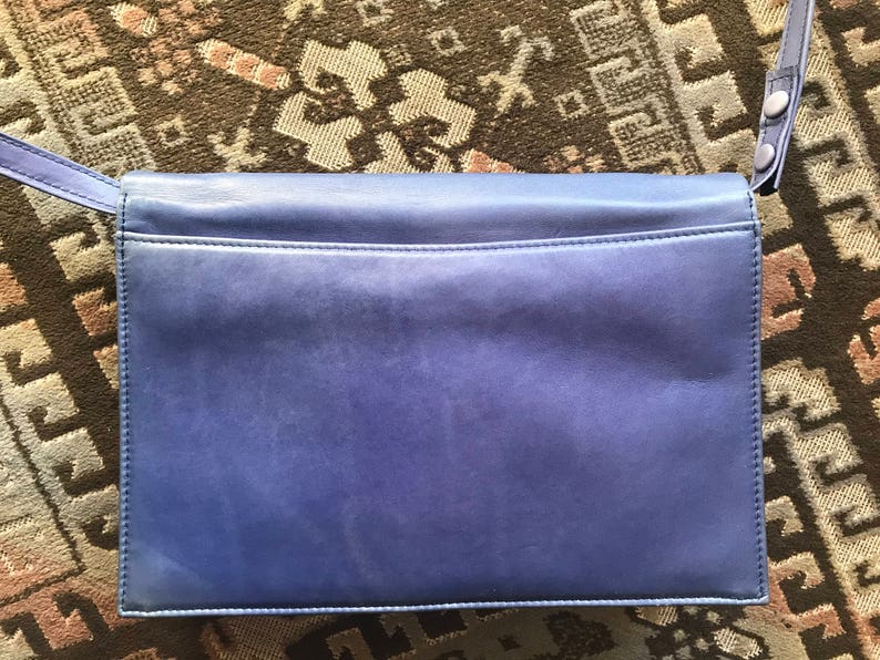 May include: A rectangular, periwinkle leather handbag with a shoulder strap. The bag features a flap closure and a subtle texture. The strap is attached with silver-toned hardware. The bag is set against a patterned, neutral-toned background.