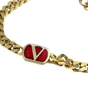 K&ouml;nnte beinhalten: Goldfarbenes Kettenarmband mit einem rechteckigen roten Emaille-Akzent, der ein goldenes "V"-Logo zeigt. Das Armband hat ein klassisches Panzerketten-Design und einen sicheren Verschluss. Ein stilvolles Accessoire.