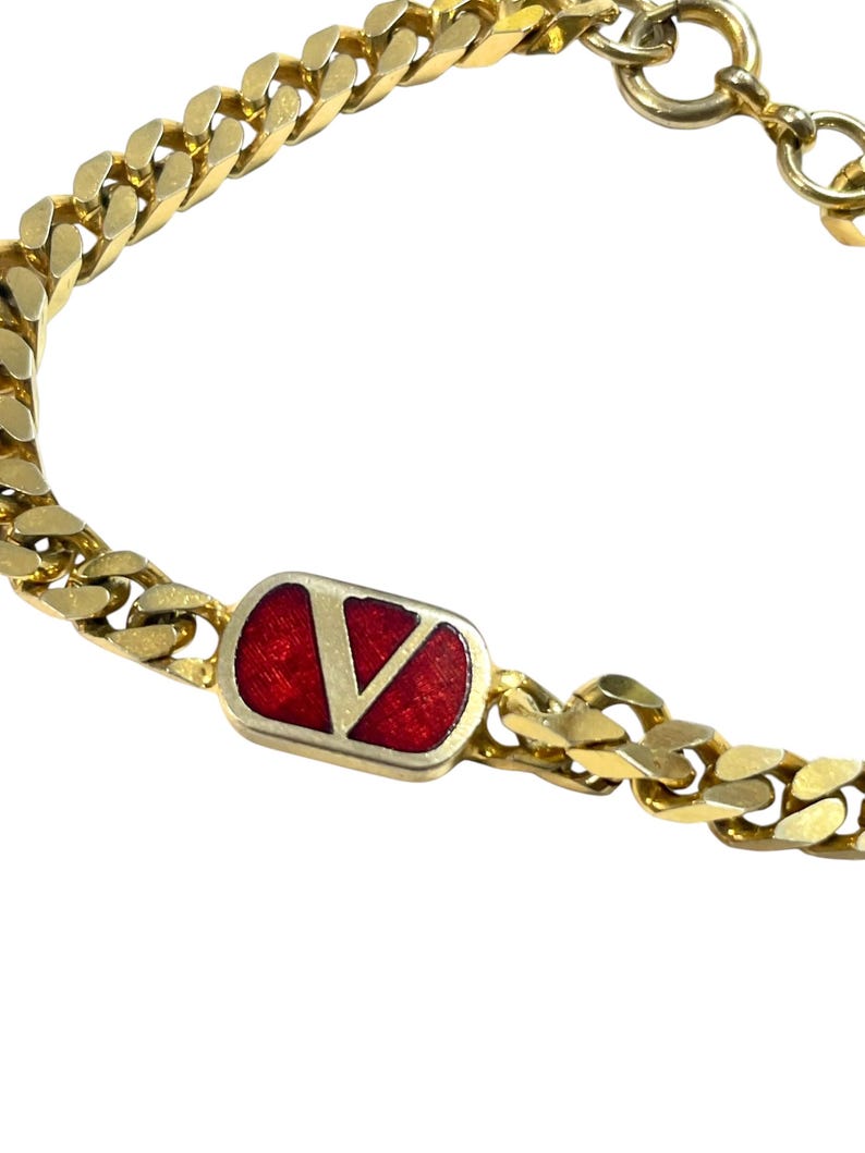K&ouml;nnte beinhalten: Goldfarbenes Kettenarmband mit einem rechteckigen roten Emaille-Akzent, der ein goldenes "V"-Logo zeigt. Das Armband hat ein klassisches Panzerketten-Design und einen sicheren Verschluss. Ein stilvolles Accessoire.