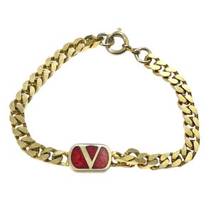 K&ouml;nnte beinhalten: Goldfarbenes Kettenarmband mit einem rechteckigen roten Emaille-Akzent und einem wei&szlig;en V-f&ouml;rmigen Logo. Das Armband hat ein klassisches Panzerketten-Design und einen sicheren Verschluss.