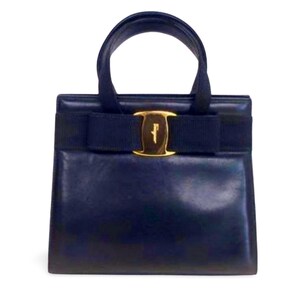 May include: A navy blue handbag with a top handle and a gold-tone clasp. The bag features a bow detail and a rectangular shape. The handbag is made of leather and has a structured design.