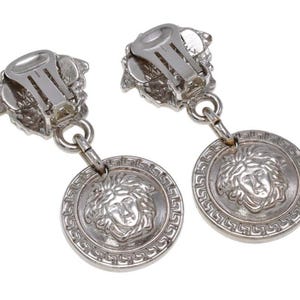 Pode incluir: Um par de brincos prateados de encaixe. Cada brinco apresenta um desenho de cabe&ccedil;a de Medusa num pendente circular, com uma borda grega. O mecanismo de encaixe &eacute; vis&iacute;vel na parte superior.