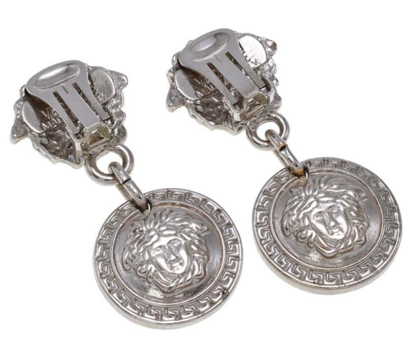 Pode incluir: Um par de brincos prateados de encaixe. Cada brinco apresenta um desenho de cabe&ccedil;a de Medusa num pendente circular, com uma borda grega. O mecanismo de encaixe &eacute; vis&iacute;vel na parte superior.