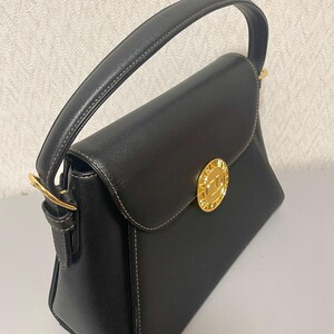 Puede incluir: Bolso de mano negro con dise&ntilde;o estructurado. Cuenta con un asa superior, cierre de solapa y herrajes dorados. El bolso tiene un emblema dorado en la parte delantera y detalles de costuras blancas. El bolso est&aacute; hecho de un material texturizado.