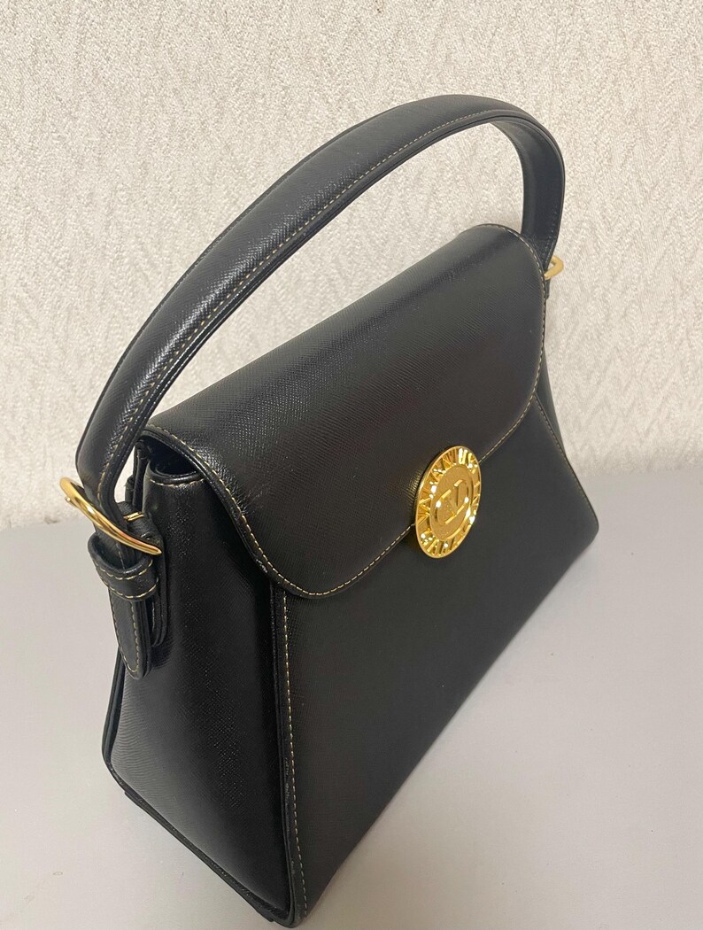 Puede incluir: Bolso de mano negro con dise&ntilde;o estructurado. Cuenta con un asa superior, cierre de solapa y herrajes dorados. El bolso tiene un emblema dorado en la parte delantera y detalles de costuras blancas. El bolso est&aacute; hecho de un material texturizado.