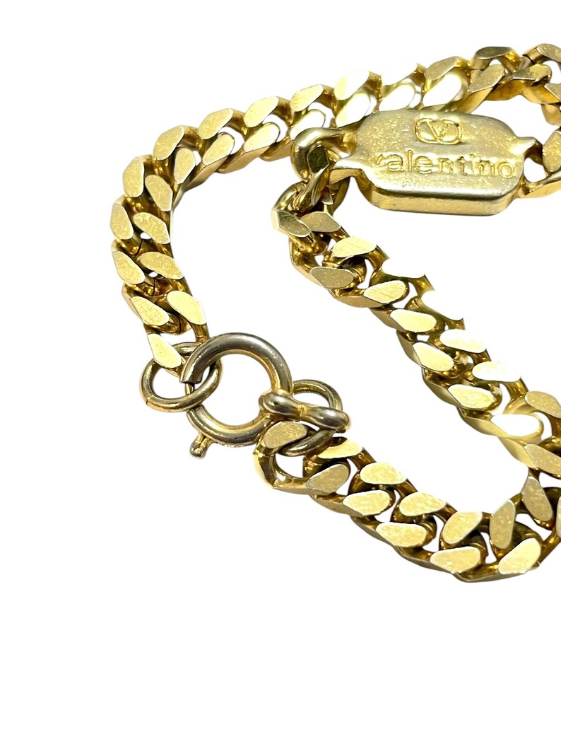 K&ouml;nnte beinhalten: Nahaufnahme einer goldfarbenen Halskette mit einem klobigen Panzerketten-Design. Die Halskette hat einen Verschluss und ein rechteckiges Etikett mit dem eingravierten Markennamen "valentino". Der Schmuck ist vor einem wei&szlig;en Hintergrund platziert.