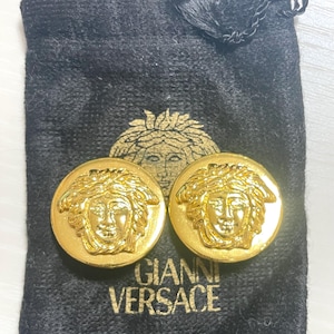 May include: A pair of gold-tone earrings featuring a raised Medusa head design. The earrings are in a black velvet pouch with the text "Gianni Versace" printed on it.
