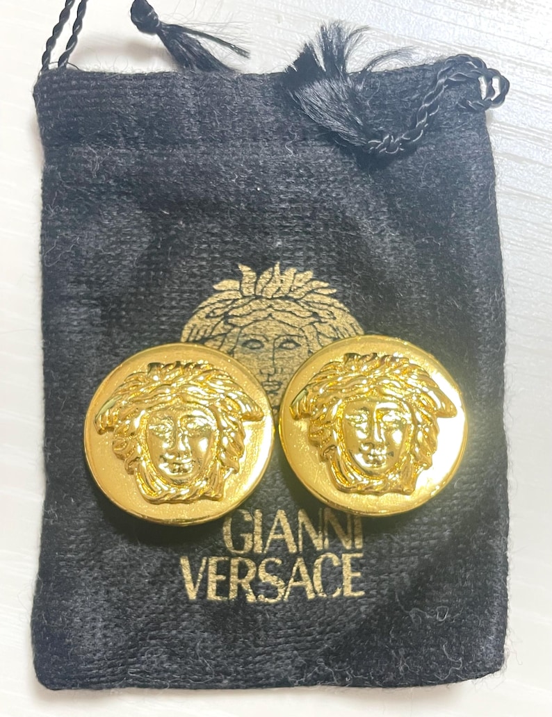 May include: A pair of gold-tone earrings featuring a raised Medusa head design. The earrings are in a black velvet pouch with the text "Gianni Versace" printed on it.