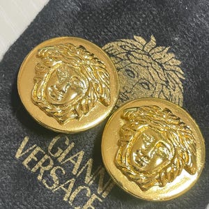 May include: A pair of gold-tone earrings featuring a raised Medusa head design. The earrings are on a black velvet background with the text "Gianni Versace" printed in gold.