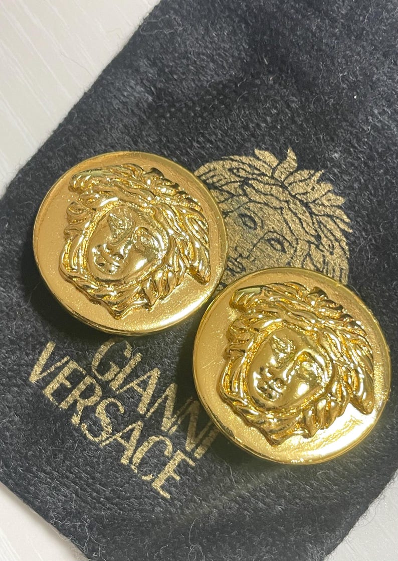 May include: A pair of gold-tone earrings featuring a raised Medusa head design. The earrings are on a black velvet background with the text "Gianni Versace" printed in gold.