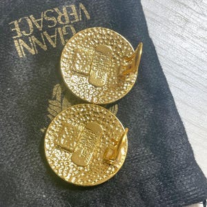 May include: A pair of gold-tone clip-on earrings with a textured circular design. The earrings feature the word "VERSACE" in a raised design.