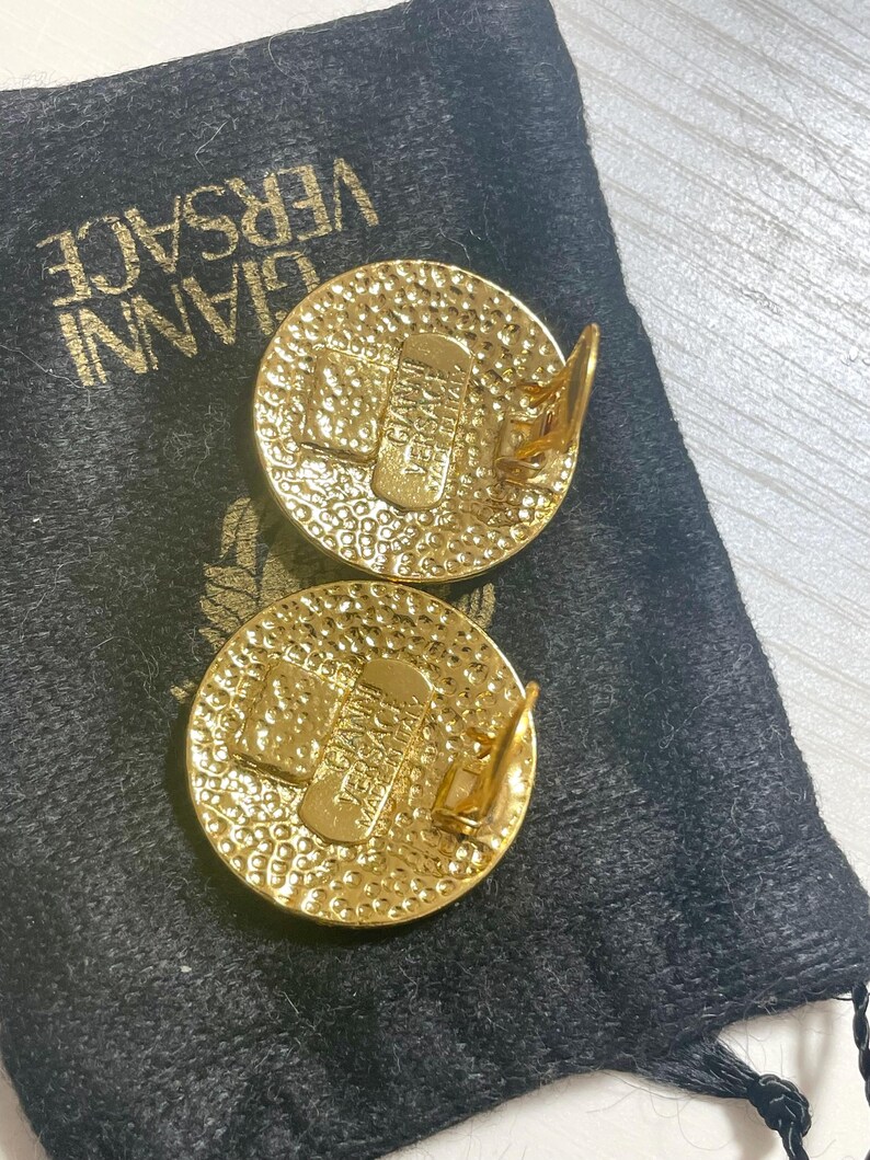 May include: A pair of gold-tone clip-on earrings with a textured circular design. The earrings feature the word "VERSACE" in a raised design.