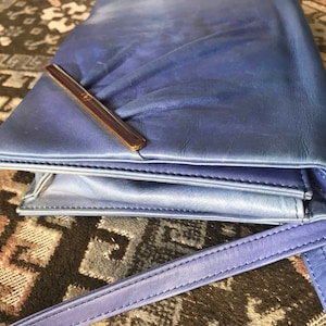 May include: A blue leather clutch bag with a shoulder strap. The bag has a rectangular shape with a flap closure and a brown metal bar. The leather has a slight sheen and a gradient effect, transitioning from a lighter blue to a darker shade.