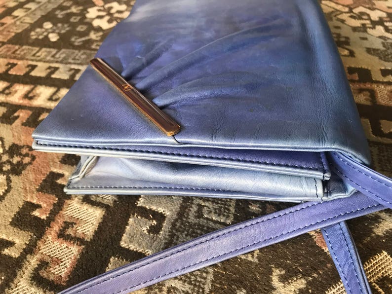 May include: A blue leather clutch bag with a shoulder strap. The bag has a rectangular shape with a flap closure and a brown metal bar. The leather has a slight sheen and a gradient effect, transitioning from a lighter blue to a darker shade.