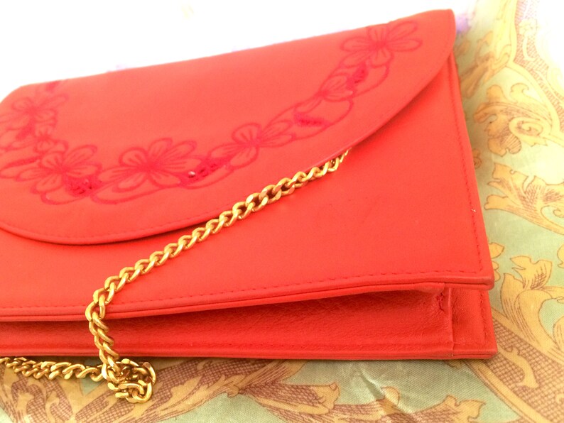 May include: A bright orange leather clutch purse with a gold chain strap and a floral design.