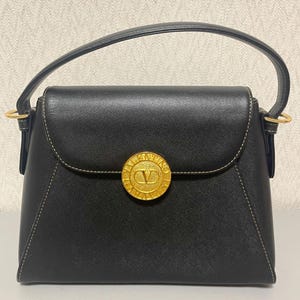Puede incluir: Bolso de mano negro con un cierre circular dorado con un logotipo 'V'. El bolso tiene una solapa superior curvada, un asa superior y herrajes dorados. Las costuras son de color beige claro.