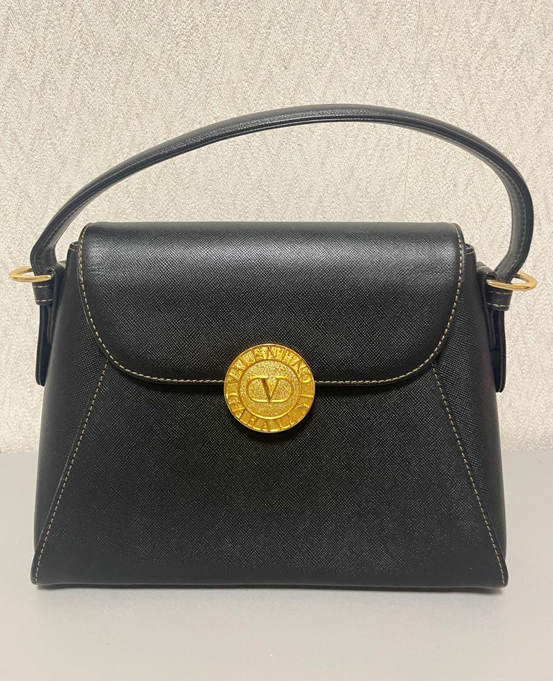 Puede incluir: Bolso de mano negro con un cierre circular dorado con un logotipo 'V'. El bolso tiene una solapa superior curvada, un asa superior y herrajes dorados. Las costuras son de color beige claro.