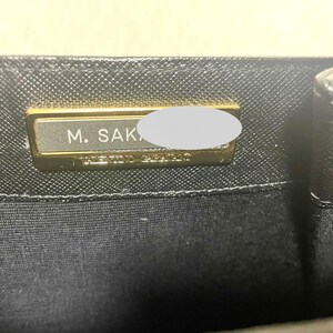 Puede incluir: Primer plano del interior de un bolso negro con una placa rectangular dorada. La placa tiene el texto "M. SAK" y "VALENTINO GARAVANI". El bolso tiene un patr&oacute;n texturizado.