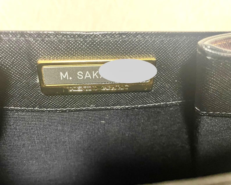 Puede incluir: Primer plano del interior de un bolso negro con una placa rectangular dorada. La placa tiene el texto "M. SAK" y "VALENTINO GARAVANI". El bolso tiene un patr&oacute;n texturizado.