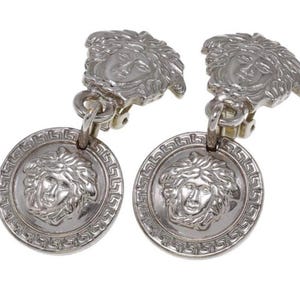 Pode incluir: Um par de brincos prateados com clip. Cada brinco apresenta um design de cabe&ccedil;a de Medusa tanto no clip como num pendente circular. O pendente tem uma borda grega. Um acess&oacute;rio de joalharia cl&aacute;ssico.