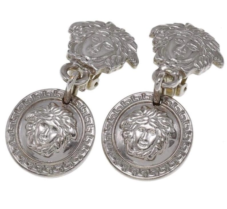 Pode incluir: Um par de brincos prateados com clip. Cada brinco apresenta um design de cabe&ccedil;a de Medusa tanto no clip como num pendente circular. O pendente tem uma borda grega. Um acess&oacute;rio de joalharia cl&aacute;ssico.