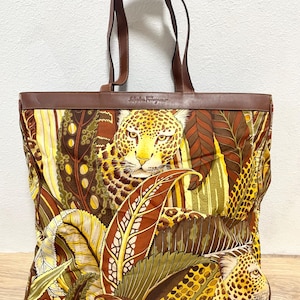 May include: A Salvatore Ferragamo tote bag featuring a vibrant jungle print with leopards and tropical leaves in shades of yellow, brown, and green. The bag has brown leather trim, a top leather band with the brand name, and two leather handles.
