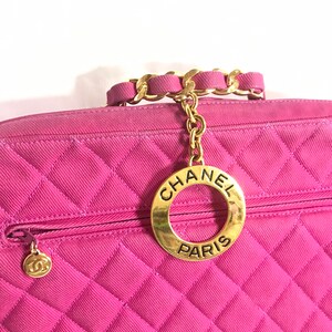 May include: A pink quilted handbag with a gold chain strap and a gold circular charm with the text "CHANEL PARIS".