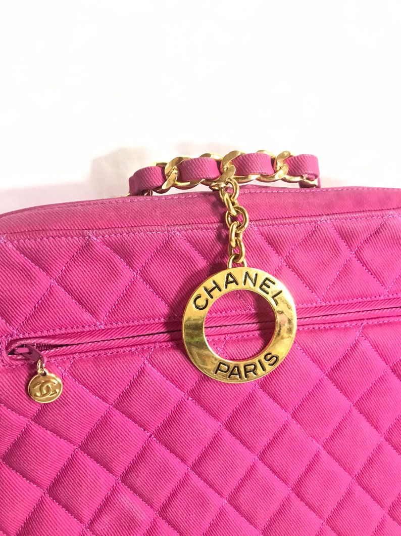 May include: A pink quilted handbag with a gold chain strap and a gold circular charm with the text "CHANEL PARIS".
