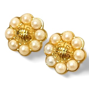 Puede incluir: Un par de pendientes de tono dorado con un diseño floral. Cada pendiente tiene una pieza central con un diseño texturizado y está rodeado por un círculo de perlas falsas.
