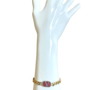 K&ouml;nnte beinhalten: Goldfarbenes Kettenarmband mit einem rechteckigen roten und silbernen Akzent. Der Akzent zeigt ein rotes 'V'-Logo. Das Armband wird auf einem wei&szlig;en Mannequin-Hand vor wei&szlig;em Hintergrund pr&auml;sentiert.