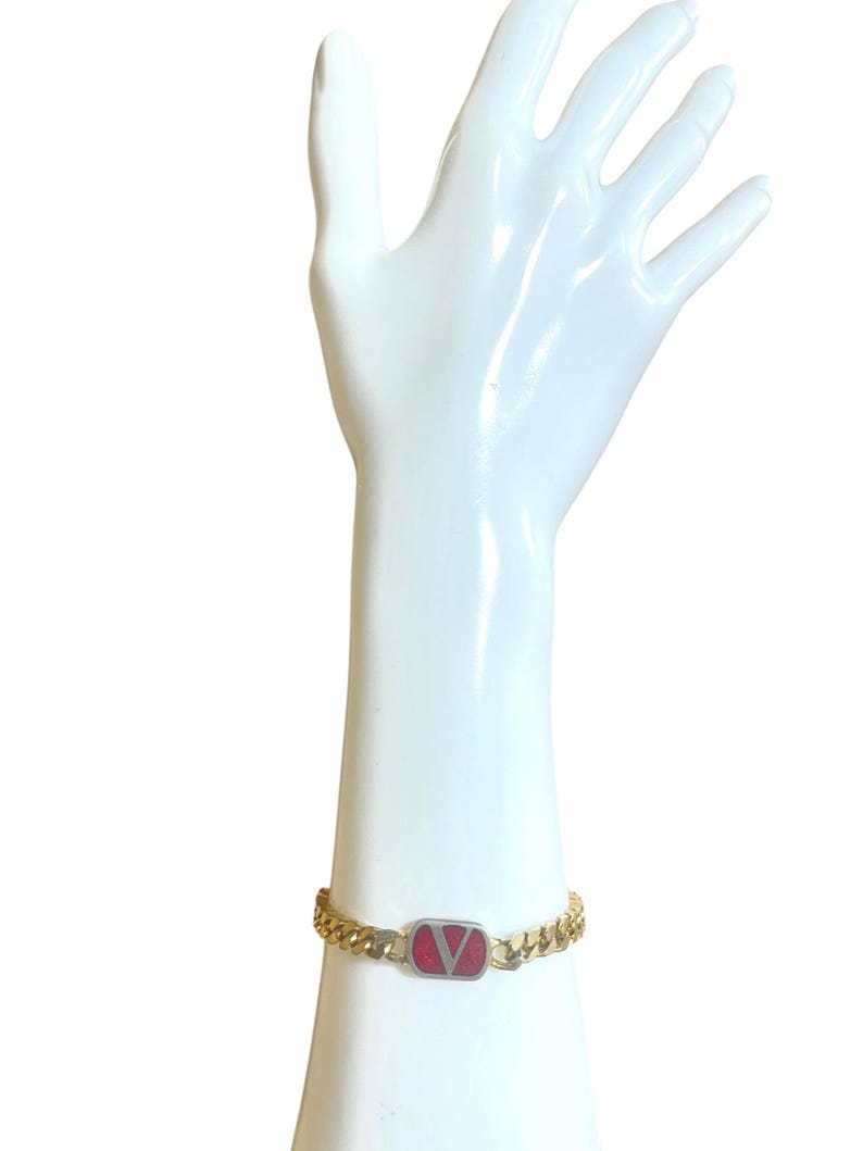 K&ouml;nnte beinhalten: Goldfarbenes Kettenarmband mit einem rechteckigen roten und silbernen Akzent. Der Akzent zeigt ein rotes 'V'-Logo. Das Armband wird auf einem wei&szlig;en Mannequin-Hand vor wei&szlig;em Hintergrund pr&auml;sentiert.