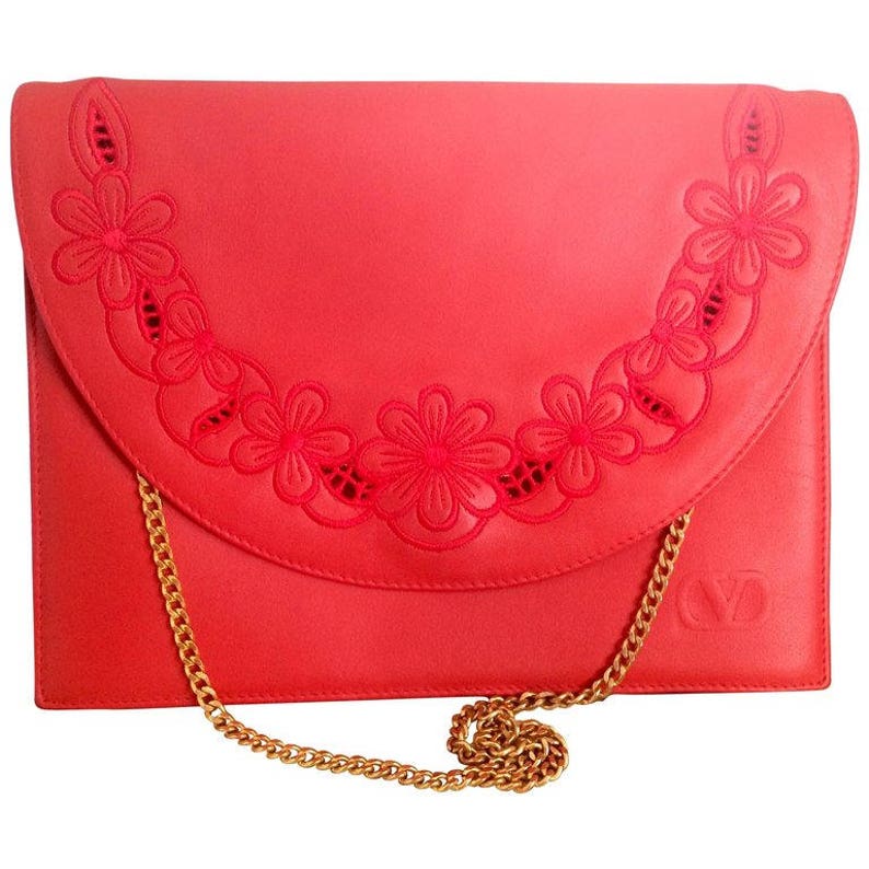 May include: A coral red leather clutch with a gold chain strap. The clutch has a floral design cut out of the leather.
