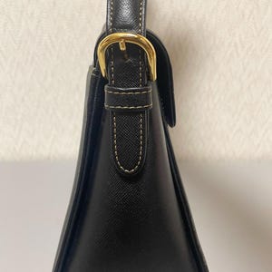 Puede incluir: Un bolso negro con una forma triangular estructurada. El bolso presenta una hebilla y una correa doradas, con detalles de costura visibles. El asa tambi&eacute;n es negra y el bolso parece estar hecho de cuero o un material similar.