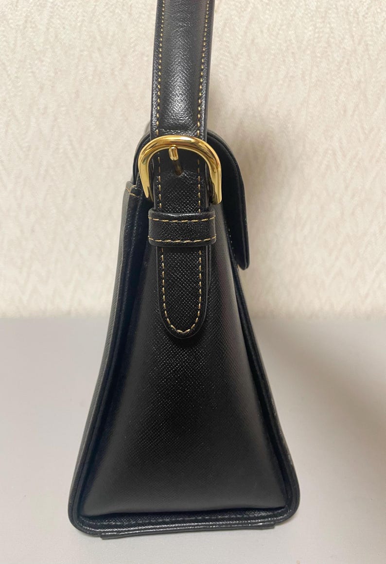 Puede incluir: Un bolso negro con una forma triangular estructurada. El bolso presenta una hebilla y una correa doradas, con detalles de costura visibles. El asa tambi&eacute;n es negra y el bolso parece estar hecho de cuero o un material similar.
