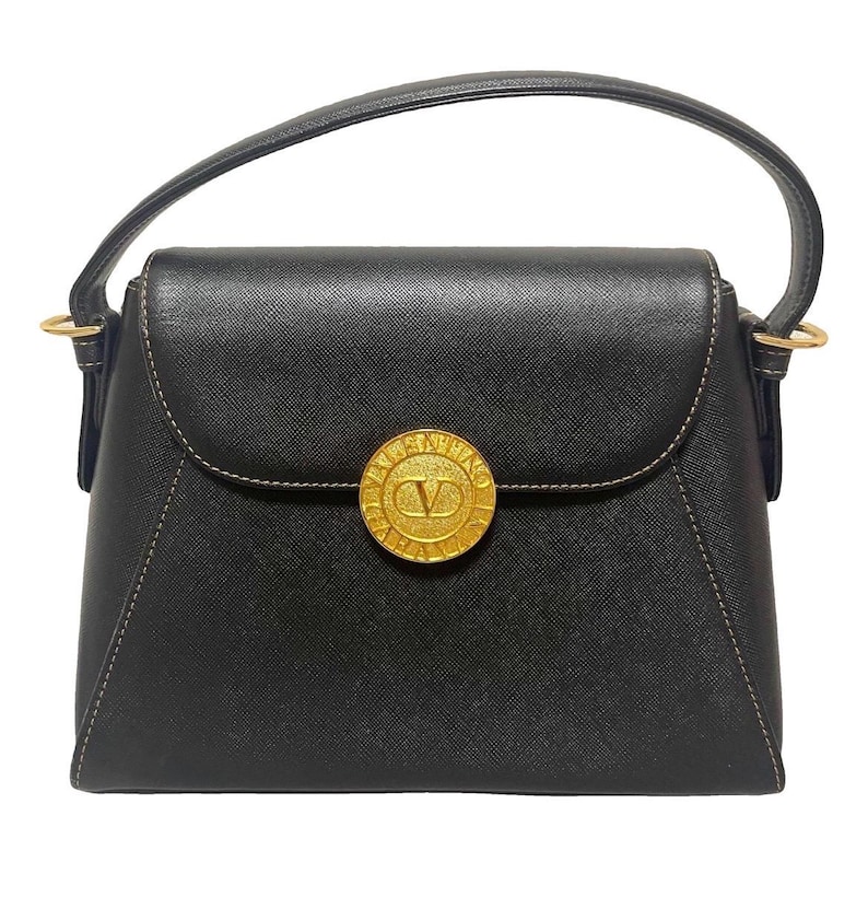 Puede incluir: Bolso de mano negro con solapa superior curvada y un emblema circular dorado. El emblema presenta la letra "V" dentro de un c&iacute;rculo. El bolso tiene un asa superior y herrajes dorados.
