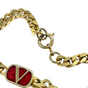 K&ouml;nnte beinhalten: Nahaufnahme eines goldfarbenen Kettenarmbands. Das Armband hat einen rechteckigen roten Emaille-Akzent mit einem goldenen "V"-Logo. Die Kettenglieder sind dick und flach. Der Verschluss ist sichtbar, und der Hintergrund ist wei&szlig;.