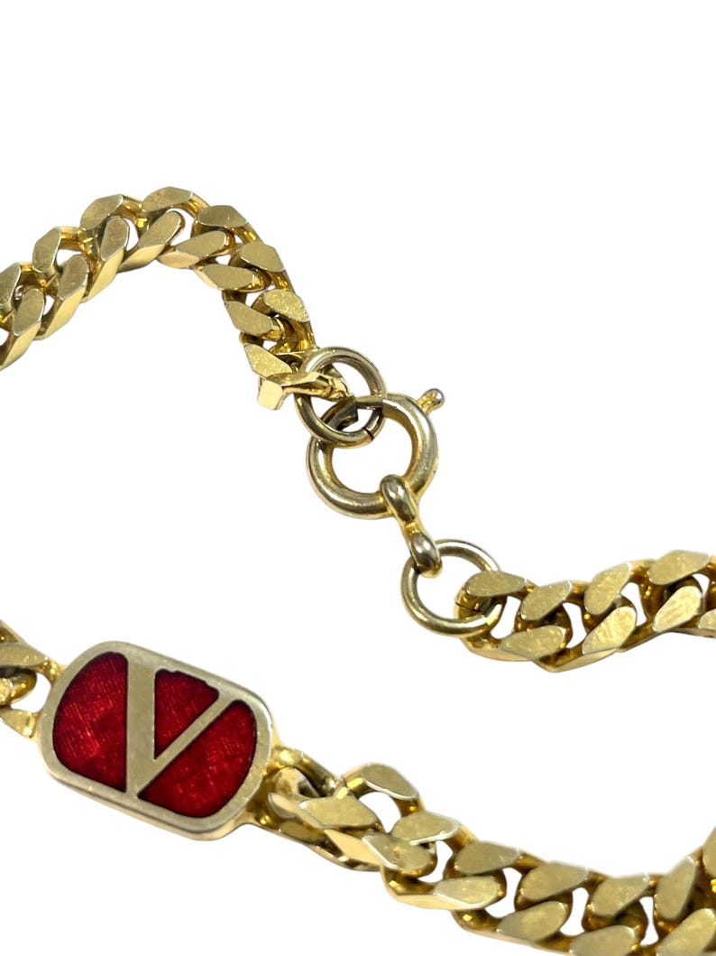 K&ouml;nnte beinhalten: Nahaufnahme eines goldfarbenen Kettenarmbands. Das Armband hat einen rechteckigen roten Emaille-Akzent mit einem goldenen "V"-Logo. Die Kettenglieder sind dick und flach. Der Verschluss ist sichtbar, und der Hintergrund ist wei&szlig;.
