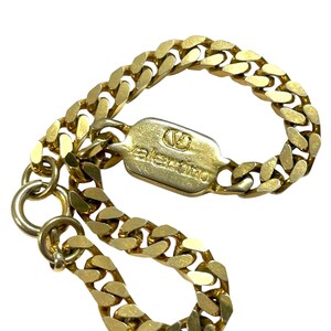 K&ouml;nnte beinhalten: Ein goldfarbenes Kettenarmband mit einem rechteckigen Verschluss. Der Verschluss ist mit dem Markennamen "Valentino" und einem Logo graviert. Die Kettenglieder sind dick und abgerundet. Das Armband wird vor wei&szlig;em Hintergrund gezeigt.
