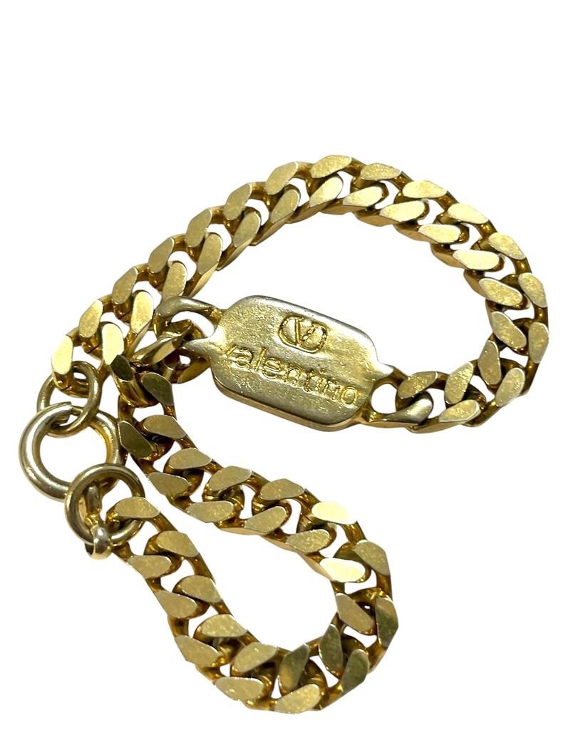 K&ouml;nnte beinhalten: Ein goldfarbenes Kettenarmband mit einem rechteckigen Verschluss. Der Verschluss ist mit dem Markennamen "Valentino" und einem Logo graviert. Die Kettenglieder sind dick und abgerundet. Das Armband wird vor wei&szlig;em Hintergrund gezeigt.