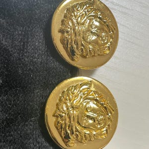 May include: A pair of gold-tone earrings featuring a raised Medusa head design. The earrings are on a black velvet background.