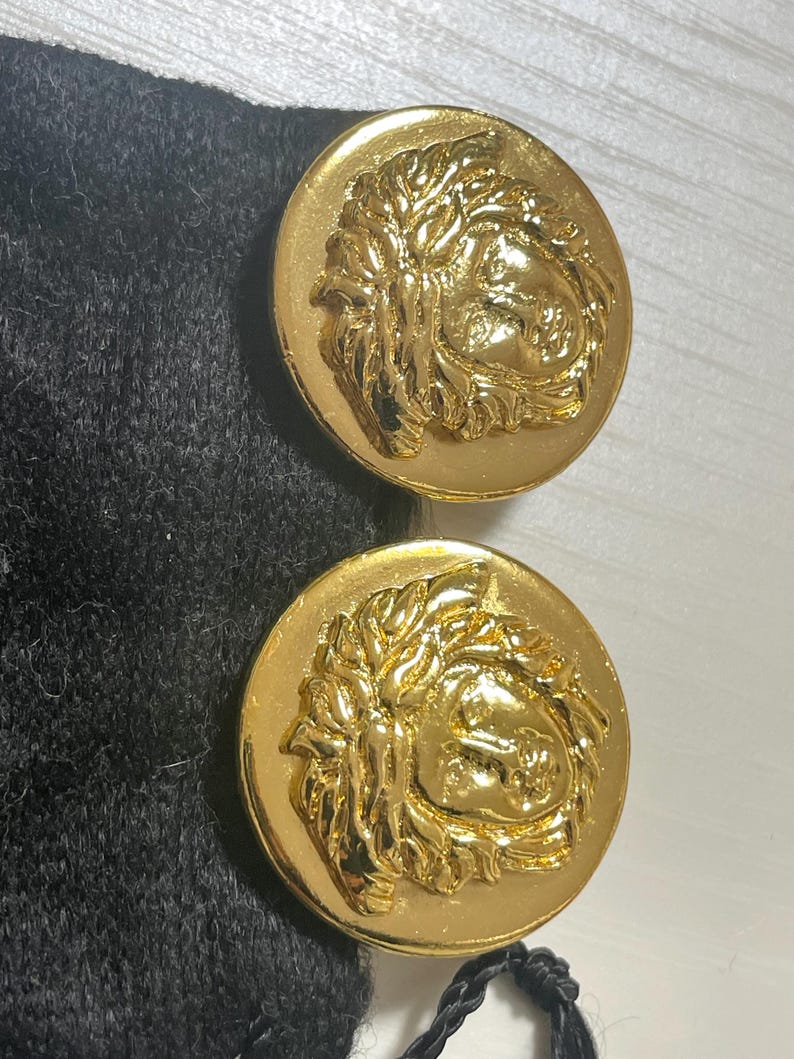 May include: A pair of gold-tone earrings featuring a raised Medusa head design. The earrings are on a black velvet background.