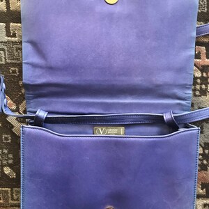 May include: A vintage royal blue leather handbag, open to show the interior. It has a gold-tone clasp and a long shoulder strap. The interior reveals a brand label. The bag is a classic design.