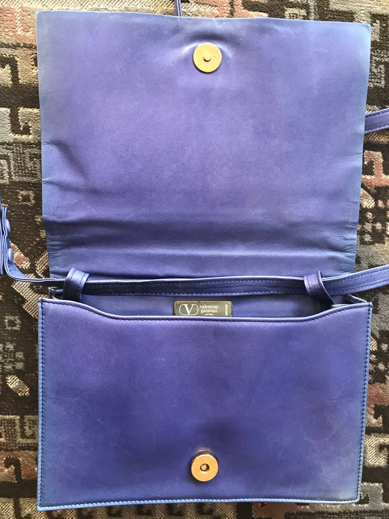 May include: A vintage royal blue leather handbag, open to show the interior. It has a gold-tone clasp and a long shoulder strap. The interior reveals a brand label. The bag is a classic design.