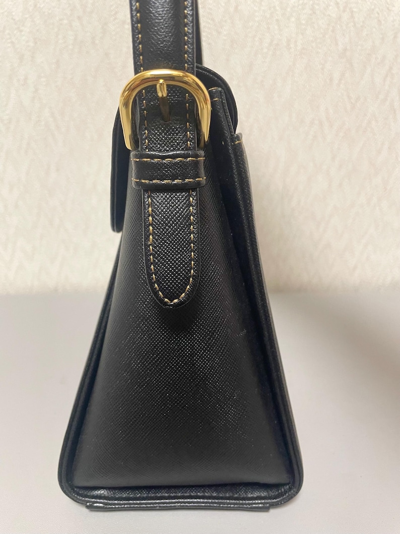 Puede incluir: Bolso de mano negro con forma trapezoidal estructurada. Cuenta con una hebilla dorada y detalles de costura. El asa es negra con costuras doradas a juego. El bolso tiene un dise&ntilde;o cl&aacute;sico y elegante.
